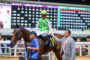 Jockey in bright green vest wearing a helmet on a brown horse being guided by handlers near a crowded railing with a large scoreboard in the background