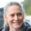 Close-up of a smiling older woman with gray hair outdoors, wearing a blue jacket.