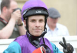 Close-up of a person wearing a purple helmet and purple-teal racing suit, looking at the camera with a neutral expression.