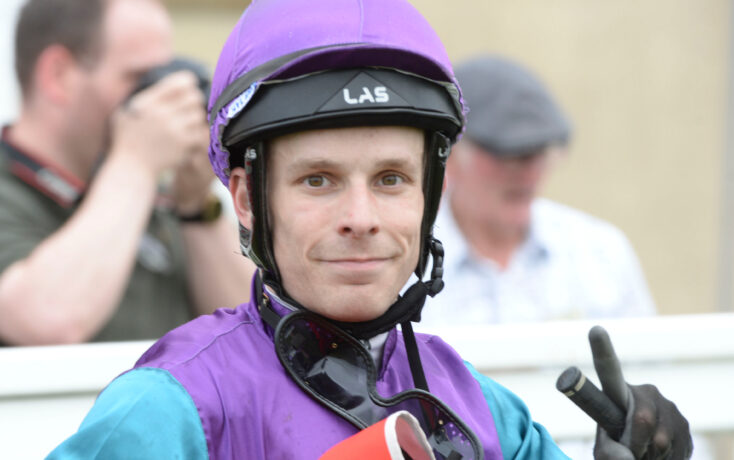 Close-up of a person wearing a purple helmet and purple-teal racing suit, looking at the camera with a neutral expression.