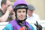 Close-up of a person wearing a purple helmet and purple-teal racing suit, looking at the camera with a neutral expression.