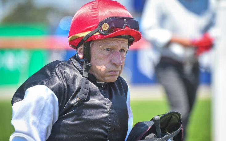 Close-up of an older jockey wearing a red helmet and goggles, black vest, white sleeves, outdoors at a race course.