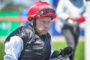 Close-up of an older jockey wearing a red helmet and goggles, black vest, white sleeves, outdoors at a race course.