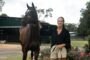 Woman in a black blouse and beige shorts stands beside a brown horse, holding its reins in a farm yard with stables in the background and blooming plants nearby.